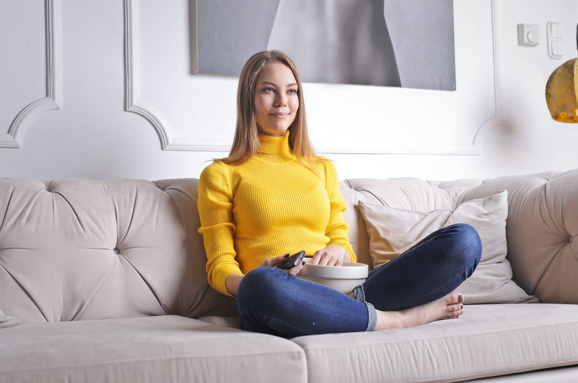 young woman in yellow jersy sitting on the sofa with popcorn on laps and remote in rand watching TV