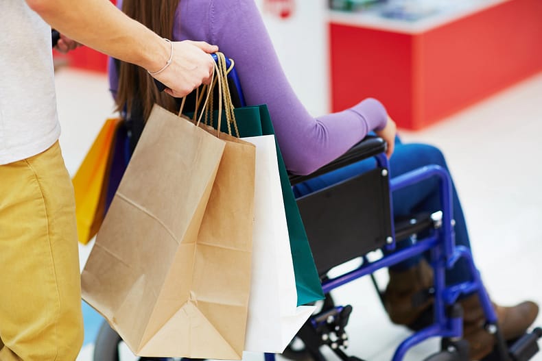 Close-up view of man with paper bags pushing her female friend in wheelchair while walking in mall